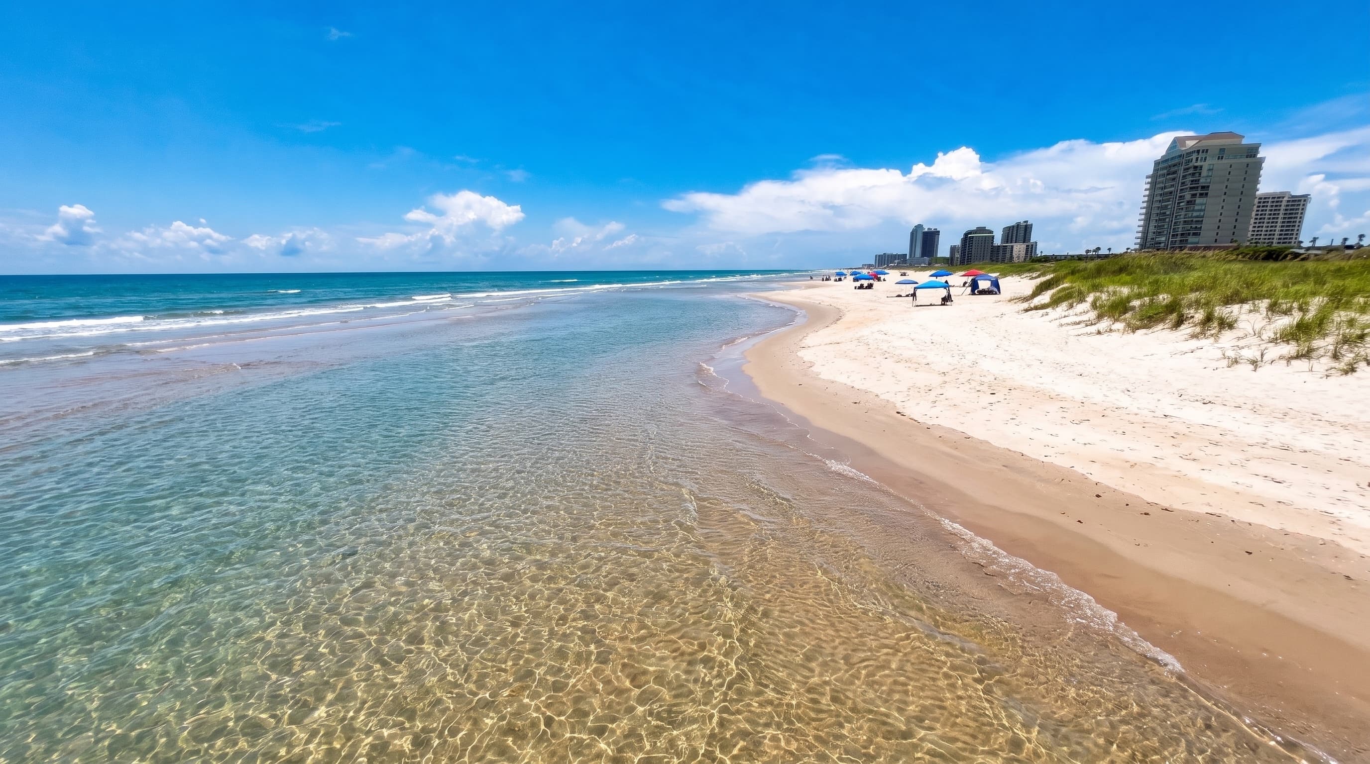 Crystal-clear, shallow low-tide water over rippled sand on South Padre Island, with condo towers along the beach